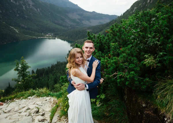 A noiva e o noivo perto do lago nas montanhas. Um casal juntos contra o pano de fundo de uma paisagem montanhosa. Morskie Oko Lake. Montanhas Tatra na Polónia . — Fotografia de Stock