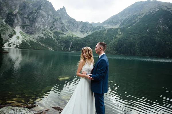 Smyslný svatební pár stojící na kamenitém břehu jezera Sea Eye v Polsku. — Stock fotografie