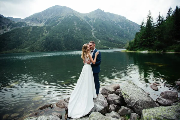 Bride with beautiful white dress and bride overlooking beautiful green mountains and lake with blue water — Stock Photo, Image