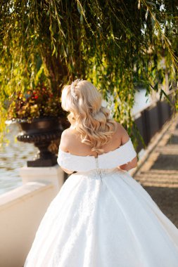 Uzun trenli beyaz gelinlikli gelin yeşil bir parkta duruyor. Arka planda dağlar ve bir göl var.