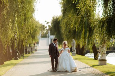 Gelin ve damat parkta birlikte yürürler. Beyaz elbiseli çekici gelin, damat koyu renkli şık bir takım giyiyor..