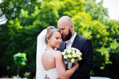 Genç ve güzel bir gelin ve kocası bir buket çiçekle bir yaz parkında dikiliyor..