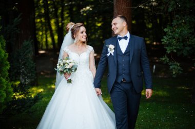 Gelin ve damat parkta birlikte yürürler. Beyaz elbiseli çekici gelin, damat koyu renkli şık bir takım giyiyor.