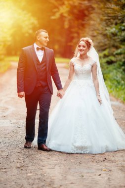 Gelin ve damat parkta birlikte yürürler. Beyaz elbiseli çekici gelin, damat koyu renkli şık bir takım giyiyor.