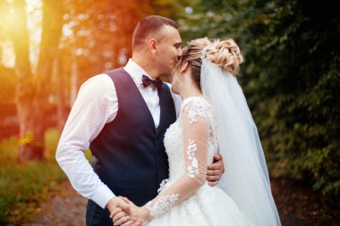 Gelin ve damat parkta birlikte yürürler. Beyaz elbiseli çekici gelin, damat koyu renkli şık bir takım giyiyor.