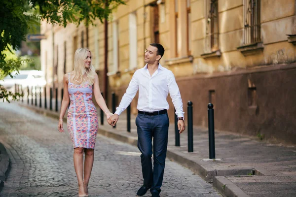可爱的夫妇在街区里散步。穿着白衬衫的黑头发男人拥抱着一位穿着漂亮衣服的金发姑娘 — 图库照片