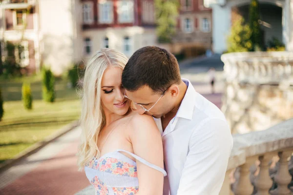 Lovely couple walking around the block. Dark-haired man in a white shirt hugging a blonde in a beautiful dress — Stock Photo, Image