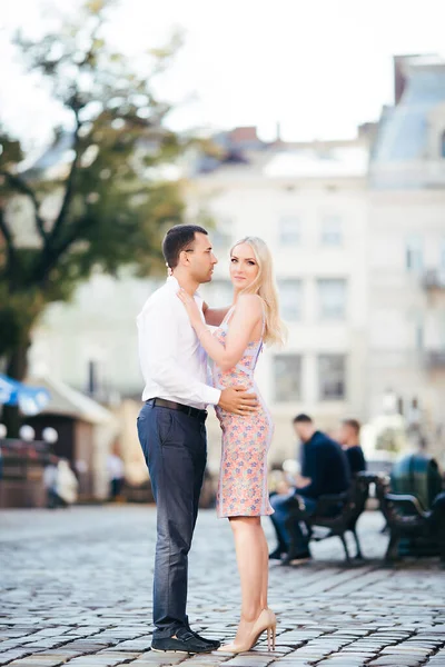 Um lindo casal a passear pelo quarteirão. Homem de cabelos escuros em uma camisa branca abraçando uma loira em um vestido bonito — Fotografia de Stock