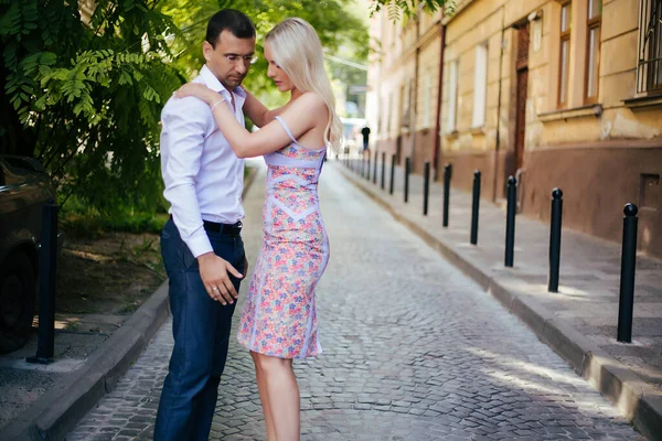 Hermosa pareja caminando por la cuadra. Hombre de pelo oscuro con una camisa blanca abrazando a una rubia en un hermoso vestido —  Fotos de Stock