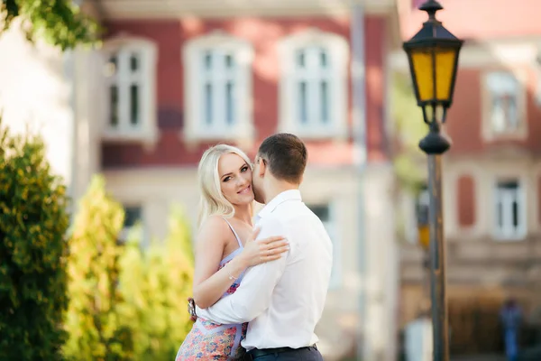 Um lindo casal a passear pelo quarteirão. Homem de cabelos escuros em uma camisa branca abraçando uma loira em um vestido bonito — Fotografia de Stock