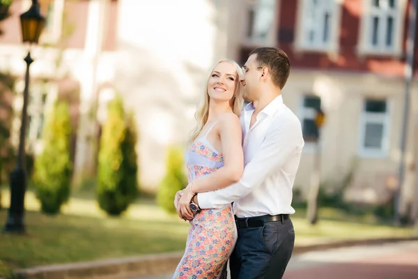 Прекрасная пара, прогуливающаяся по кварталу. Темноволосый мужчина в белой рубашке обнимает блондинку в красивом платье — стоковое фото