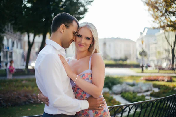 Um lindo casal a passear pelo quarteirão. Homem de cabelos escuros em uma camisa branca abraçando uma loira em um vestido bonito — Fotografia de Stock