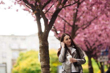 Kumral kız ceketli ve kot pantolonlu. Şehrin dışında, çiçek açan sakura ağaçlarının arka planında.