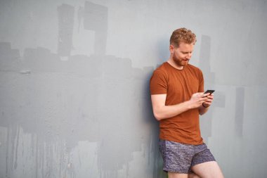 Genç adam gri renkli bir duvarın yanında akıllı telefon kullanıyor.