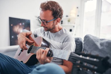 Adam oturma odasında akustik gitar çalıyor..
