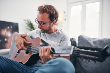 Adam oturma odasında akustik gitar çalıyor..