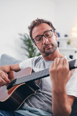 Adam oturma odasında akustik gitar çalıyor..