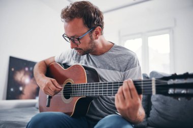 Adam oturma odasında akustik gitar çalıyor..