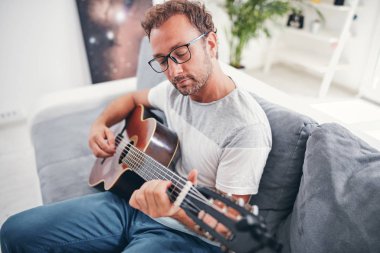 Adam oturma odasında akustik gitar çalıyor..