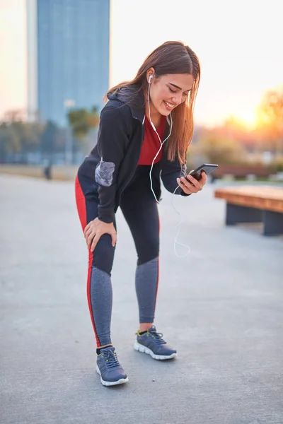 Modern young woman with cellphone making pause during jogging / — Zdjęcie stockowe