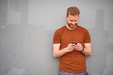 Genç adam gri renkli bir duvarın yanında akıllı telefon kullanıyor.