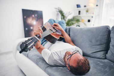 Adam oturma odasında akustik gitar çalıyor..