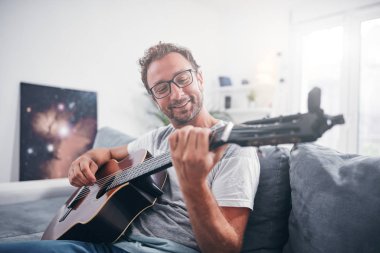 Adam oturma odasında akustik gitar çalıyor..