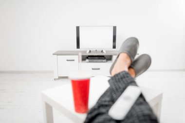 Gazoz içen ve masanın üstünde bacaklarıyla televizyona bakan bir adam.