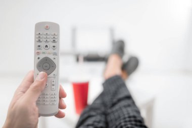 Gazoz içen ve masanın üstünde bacaklarıyla televizyona bakan bir adam.