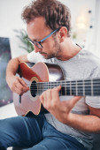 Muž hraje na akustickou kytaru v obývacím pokoji.