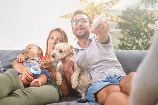 Young parents watching TV with baby boy and a dog.
