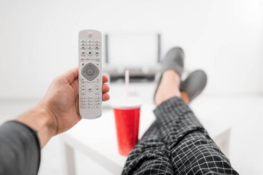Gazoz içen ve masanın üstünde bacaklarıyla televizyona bakan bir adam.
