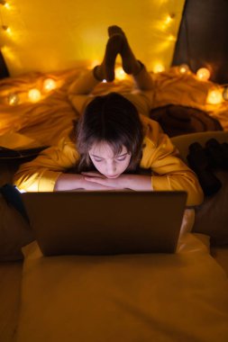 Oturma odasındaki ev yapımı çadırının altında dizüstü bilgisayar kullanan 11 yaşında bir kız..