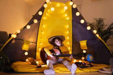 Oturma odasındaki çadırın altında akustik gitar çalan küçük kız..