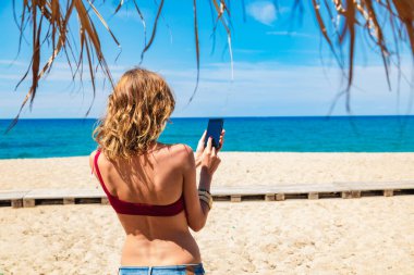Genç bir kadın tropikal kumsalda cep telefonu kullanıyor..