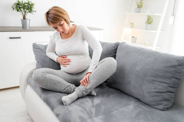 Беременная уставшая измученная женщина с проблемами с желудком, ногой и болью дома на диване, болеющая
.