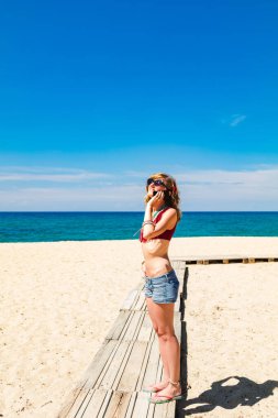 Genç bir kadın tropikal kumsalda cep telefonu kullanıyor..