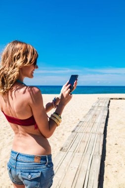 Genç bir kadın tropikal kumsalda cep telefonu kullanıyor..