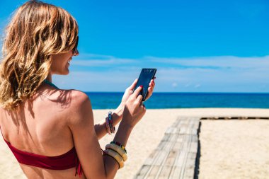 Genç bir kadın tropikal kumsalda cep telefonu kullanıyor..