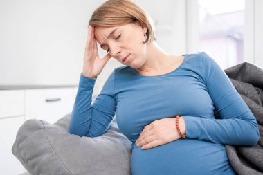 Pregnant tired exhausted woman with stomach and head issues at home on a couch, being sick.