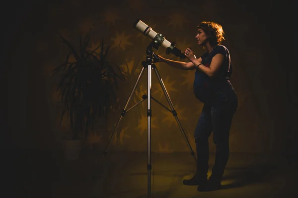 Pregnant woman looking at the stars through a telescope- astrology concept, horoscope predictions about the baby.