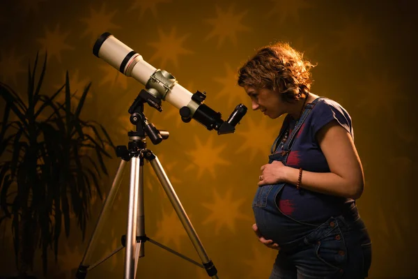 Pregnant woman looking at the stars through a telescope- astrology concept, horoscope predictions about the baby.