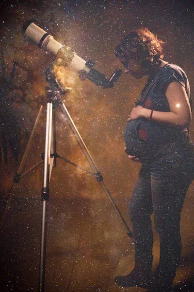 Pregnant woman looking at the stars through a telescope- astrology concept, horoscope predictions about the baby.