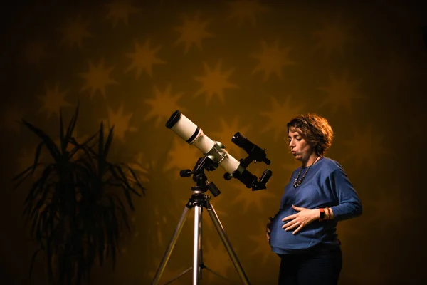 Pregnant woman looking at the stars through a telescope- astrology concept, horoscope predictions about the baby.