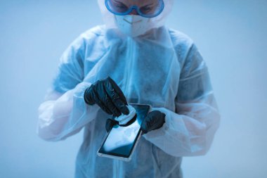 Tıp doktoru, akıllı telefon dezenfeksiyonu için antiseptik doku taşıyan virüs ve hastalıklardan korunmak için tam vücut kıyafeti giyen bilim adamı..