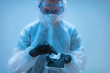 Tıp doktoru, akıllı telefon dezenfeksiyonu için antiseptik doku taşıyan virüs ve hastalıklardan korunmak için tam vücut kıyafeti giyen bilim adamı..