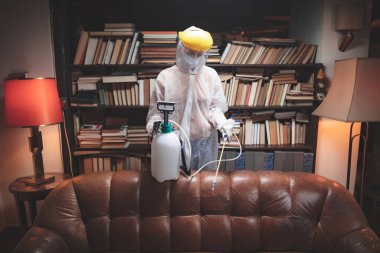 Koruyucu antiviral maskeli, kimyasal dezenfekte spreyli ev ve mobilyaları dezenfekte eden kişi..