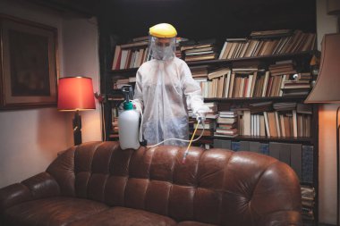 Koruyucu antiviral maskeli, kimyasal dezenfekte spreyli ev ve mobilyaları dezenfekte eden kişi..