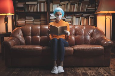 Koruyucu antiviral maskeli bir kadın evde karantinada oturuyor ve kitap okuyor..