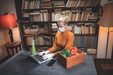 Virüs ve patojenlere karşı koruyucu maskeli bir kadın evde dizüstü bilgisayar kullanıyor, internetten yiyecek, yiyecek ve erzak sipariş ediyor..